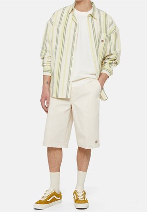 Homme portant une chemise crème rayée avec boutons, un t-shirt blanc, un short beige, des chaussettes blanches et des baskets jaune moutarde, debout avec une main dans la poche.