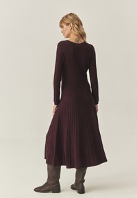 Garā piedurkņu ribota kleita dziļi bordo krāsā ar pieguļošu augšdaļu un platām plisētām svārkiem, valkāta ar brūniem ceļa garuma zābakiem.