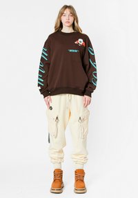 Sudadera marrón con detalles turquesa y bordados, combinada con pantalones cargo crema con cremalleras y grandes bolsillos, y botas marrones.