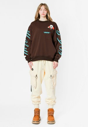 Sudadera marrón con detalles turquesa y bordados, combinada con pantalones cargo crema con cremalleras y grandes bolsillos, y botas marrones.