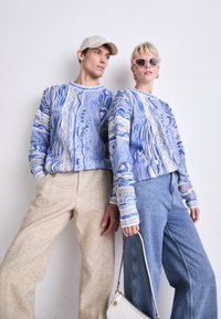 Des pulls bleus à motifs avec des designs complexes, associés à un pantalon large beige et bleu clair. La femme porte un sac à main blanc.