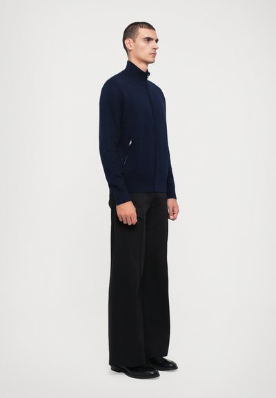 Sudadera de marina con cremallera y cuello alto, con bolsillos laterales con cremallera. Combinada con pantalones de pierna ancha negros y zapatos negros. Textura suave.