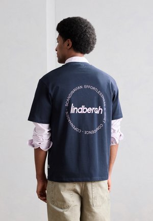 Mężczyzna z fryzurą afro, ubrany w granatowy T-shirt na podwiniętej, jasnoróżowej koszuli, beżowe spodnie, stoi na szarym tle.