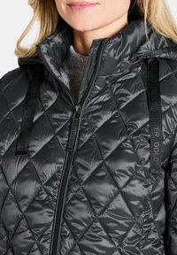 Veste matelassée noire en duvet avec une finition brillante, col montant et fermeture éclair. Comprend des bretelles avec logo sur les épaules.