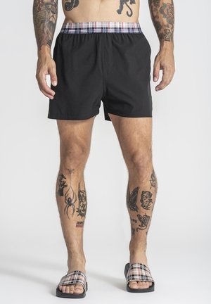 Homme portant un short noir avec une ceinture à carreaux et des sandales à glissière assorties à carreaux, affichant plusieurs tatouages sur les jambes et les bras.