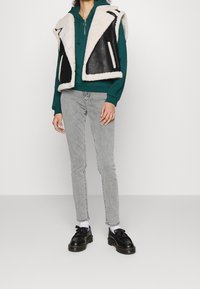 Chaleco de borreguito negro y crema sobre una sudadera verde, combinado con jeans ajustados grises y zapatos de cordones negros con calcetines blancos.
