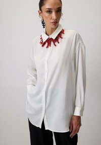 Witte blouse met lange mouwen en een knoopsluiting aan de voorkant, voorzien van een rode, koraalachtige statementkraag en glad, licht materiaal.