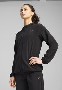 Νεαρή γυναίκα με μαύρη φόρμα Puma με ζακέτα με φερμουάρ ποζάρει με το χέρι στη μέση μπροστά σε απλό γκρι φόντο.