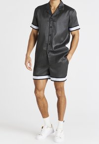 Chemise noire à manches courtes avec un motif subtil, accents blancs sur les poignets, et short assorti. Complété par des baskets blanches et des chaussettes montantes.