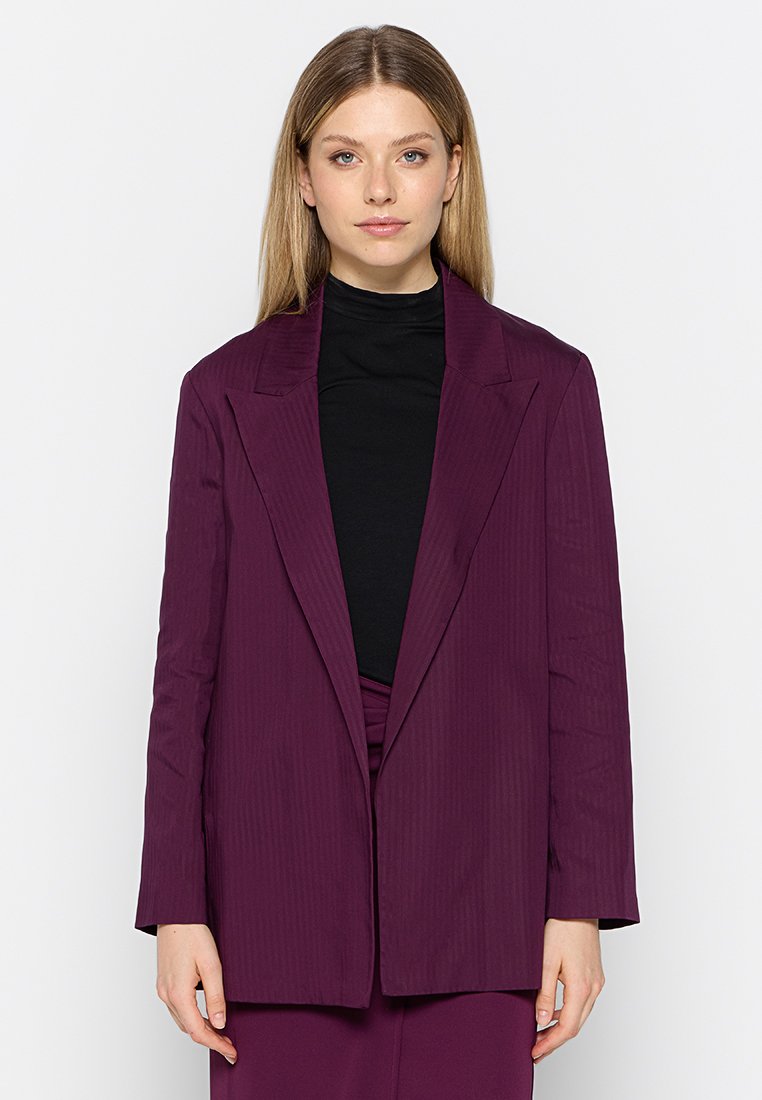 Blazer couleur prune avec un col à revers, des manches longues et une coupe ample. Tissu texturé avec des rayures verticales, porté sur un haut noir.