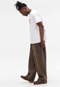 White cotton t-shirt with red graphic print on the front and sleeve, paired with brown wide-leg trousers and brown footwear.