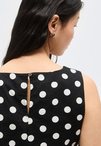 Γυναίκα με μακριά μαύρα μαλλιά, που φοράει αμάνικο μαύρο φόρεμα με μεγάλα λευκά πουά και μικρό κουμπί στο πίσω μέρος στο άνω μέρος.