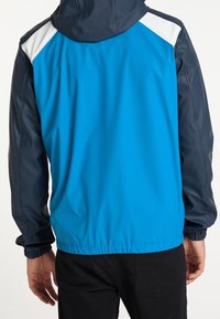 Blå och marinblå vindjacka med elastiska ärmar, huva och vita accentpaneler på axlarna. Slätt, vattentätt material.