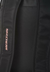 Mochila negra con textura suave, con un destacado logo rosa de "SKECHERS" en un lateral y correas acolchadas para mayor comodidad.