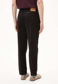 Zwarte katoenen broek met rechte snit, achterzakken en een bruin lederen logo op het tailleband. Draagt beige slippers.