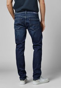 Tamsiai mėlynos džinsinės kelnės su siauru siluetu, turinčios dvi užpakalines kišenes ir geltoną siuvinėjimą. Dėvimos su baltais sportbačiais ir juoda marškinėliais.