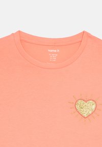 Κοραλλί βαμβακερό t-shirt με στρογγυλή λαιμόκοψη, διακοσμημένο με μια λαμπερή χρυσή καρδιά με ηλιακές ακτίνες. Ετικέτα μεγέθους ορατή στο εσωτερικό.