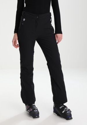 Femme portant un pantalon de ski noir imperméable et des bottes de ski noires, debout sur un fond blanc uni.