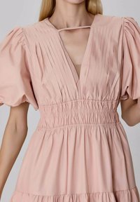 Robe à manches bouffantes en rose doux, présentant des plis verticaux sur le buste, un décolleté en V profond et une taille élastique.