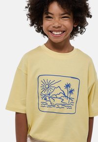 T-shirt jaune à manches courtes avec un motif bleu représentant un soleil, des montagnes et des palmiers dans un cadre rectangulaire. Tissu en coton doux.