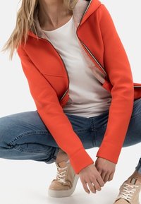 Eine Person trägt einen orangefarbenen Reißverschluss-Hoodie, ein weißes T-Shirt, blaue Jeans und beigefarbene Sneaker. Sie hockt mit den Händen nahe den Füßen.