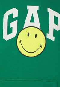 Sudadera verde con grandes letras blancas que forman "GAP" encima de un gráfico de cara sonriente amarilla con contornos negros y rasgos faciales simples.