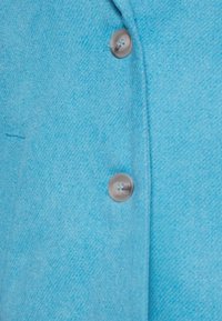 Tissu en laine bleu clair avec deux boutons ronds gris cousus verticalement sur un manteau ou une veste.