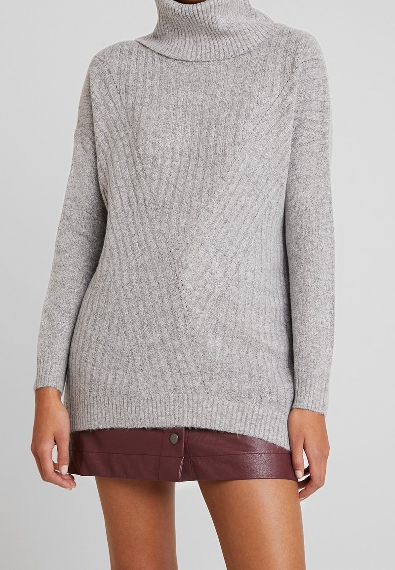 Maglione a collo alto grigio chiaro in maglia con texture a coste e design diagonale. Maniche lunghe, vestibilità rilassata e tessuto morbido. Abbinato a una gonna bordeaux.
