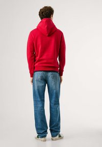 Rode hoodie met een capuchon en geribbelde manchetten, gecombineerd met lichtblauwe jeans met achterzakken en logo-detail. Sneakers met groene accenten.