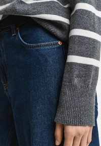Un pull rayé gris avec un ourlet côtelé, porté sur un jean en denim bleu foncé présentant des détails en rivets cuivrés et une texture lisse et soyeuse.