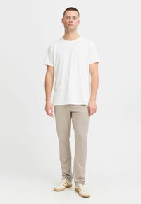 Camiseta blanca de algodón combinada con pantalones beige ajustados. Los zapatos son de color crema con franjas marrón claro. El modelo se encuentra de pie sobre un fondo claro y liso.