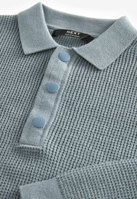 Polo shirt gris texturé avec un col, trois boutons bleu clair et un design en tricot gaufré. Étiquette de la marque visible à l'intérieur du col.