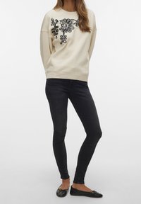 Sudadera crema con bordado floral negro, corte relajado, mangas largas, combinada con jeans skinny negros y bailarinas negras.
