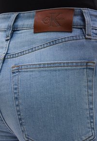 Svijetloplave traperice s kožnim zakrpom s logotipom Calvin Klein, vidljivim šavovima i dva stražnja džepa. Tekstura je glatka sa blago izblijedjelim izgledom.