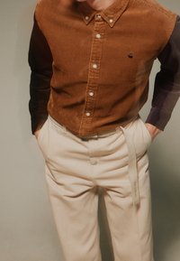 Camisa de botones de pana marrón con mangas de rayas, combinada con pantalones beiges que tienen un cinturón de tela y bolsillos laterales. Sin patrones notables.