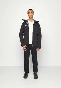 Νεαρός άνδρας στέκεται μπροστά σε ένα απλό φόντο, φορώντας ένα μαύρο μπουφάν, λευκό μπλουζάκι, μαύρο παντελόνι και μαύρα παπούτσια, κοιτάζοντας μπροστά.