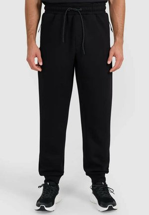 Mężczyzna noszący czarne joggery ze sznurkiem, zapinanymi na zamek bocznymi kieszeniami oraz czarno-białe buty sportowe, stojący na jasnym tle.