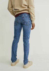 Jean en denim bleu à coupe droite, avec deux poches arrière et des coutures visibles. Porté avec un sweat-shirt beige et des chaussures claires.