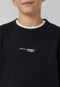 Bambino che indossa un felpa nera con la scritta bianca "ARRIVO 14:00" barrata e "cancellato" scritto sopra in font corsivo.