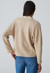 Maglione beige a trama con collo alto e maniche lunghe, caratterizzato da una cucitura sulla schiena, abbinato a jeans blu in denim.