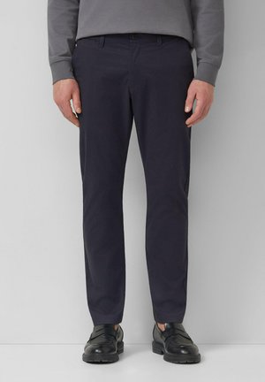 Man draagt marineblauwe slim-fit broek, grijs shirt met lange mouwen en zwarte leren nette schoenen, staand tegen een effen achtergrond.