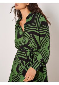Grün und schwarz gemustertes Kleid mit geometrischem Design, mit knöpfbarem Kragen, langen Ärmeln und gebundenem Taillenbund. Weicher Stoff mit glatter Textur.