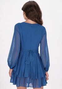 Rochie albastră cu mâneci lungi transparente și un corset ajustat. Prezintă o fustă în straturi, puncte texturate și un detaliu cu talie legat.