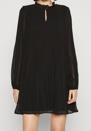 Femme portant une robe plissée à manches longues noires avec un col à volants et une petite fente au niveau du décolleté.