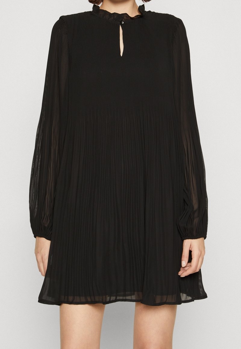 Femme portant une robe plissée à manches longues noires avec un col à volants et une petite fente au niveau du décolleté.