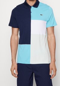 Koszulka polo z krótkim rękawem, wyróżniająca się kolorowym wzorem w odcieniach granatu, jasnoniebieskim, białym i szarym. Materiał jest gładki, z kołnierzykiem zapinanym na guziki.