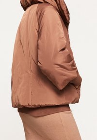 Ruskea pehmustettu takki hupulla, jossa on sileä pinta ja uurteet hihansuissa ja helmassa. Rakenteellinen, ylisuuri muotoilu.