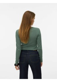 Zielony top z długim rękawem wykonany z prążkowanej tkaniny, zestawiony z ciemnoniebieskimi, wysokim stanem jeansami z tylnymi kieszeniami i ozdobnymi, złotymi szwami.
