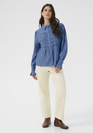 Blå skjorte med knaplukning og smockdetaljer foran, lange ærmer og puffede skuldre, parret med cremede bukser og brune ankelstøvler.