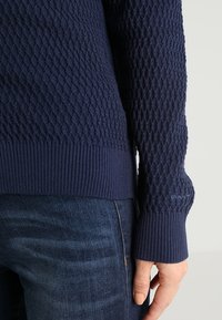 Suéter azul marino con patrón de trenzado en diamante texturizado y puños acanalados. Presenta un ajuste ceñido y una marca mínima en la manga.